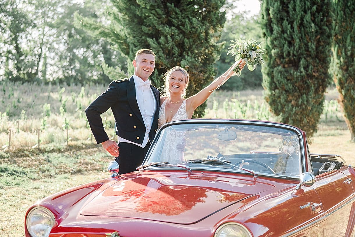 les mariés sont à coté de leur voiture de collection pour leur photos de couple et la mariée lève son bouquet de fleur au chateau de birot à Beguey